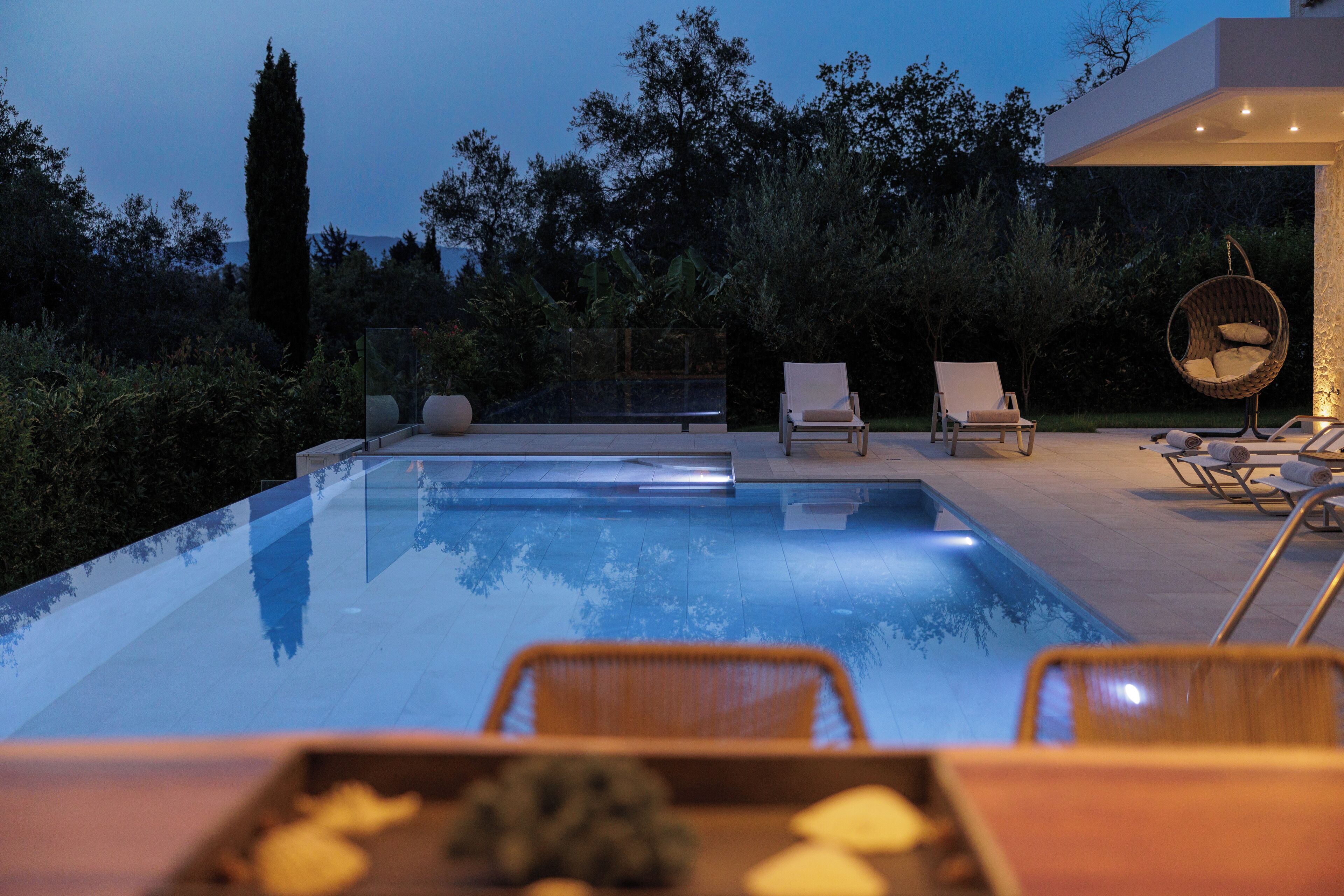A modern outdoor pool area with lounge chairs and a hanging chair, surrounded by greenery.