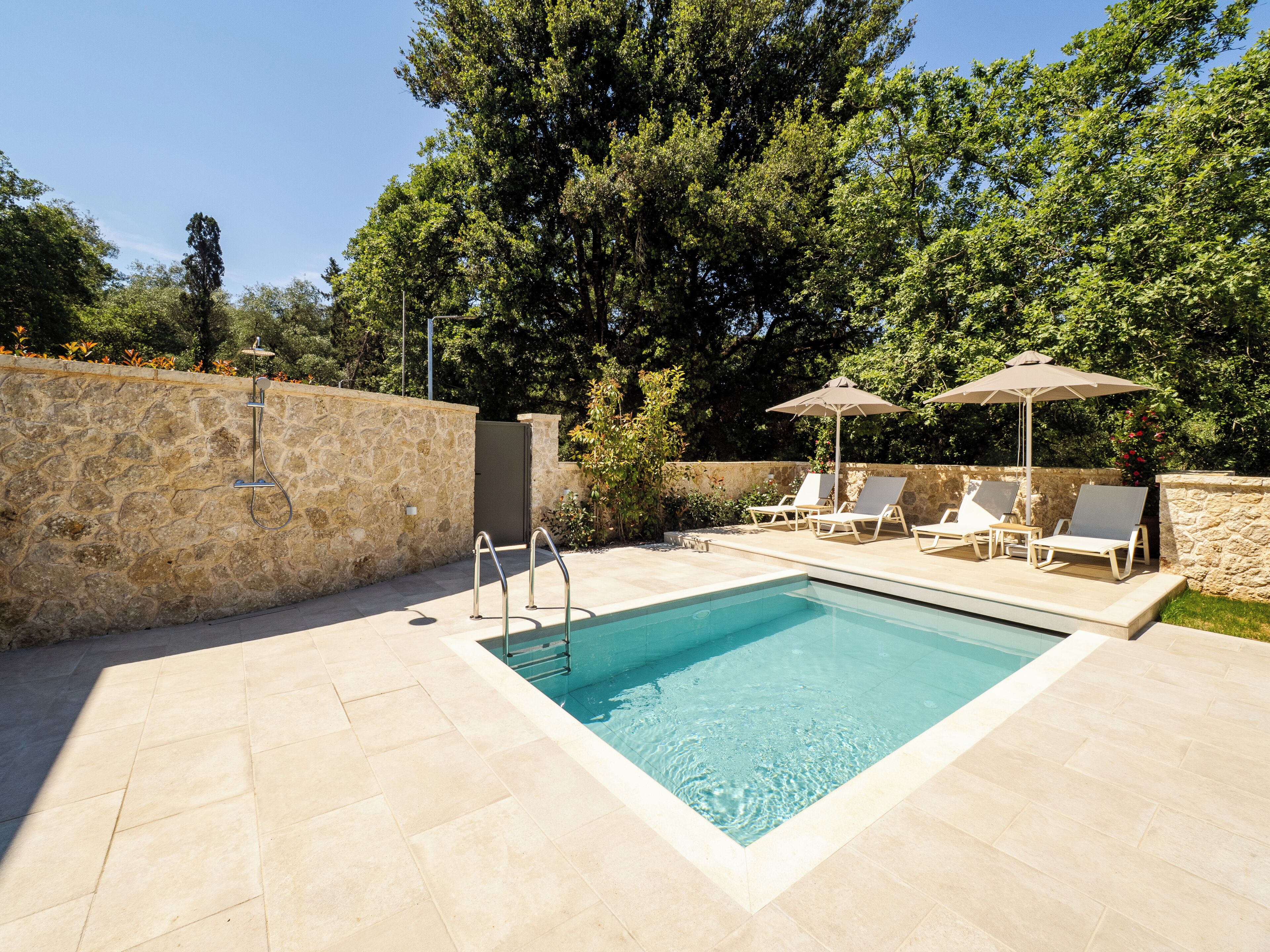 Una piscina con muro de piedra, ducha metálica y sillones de descanso con sombrillas.