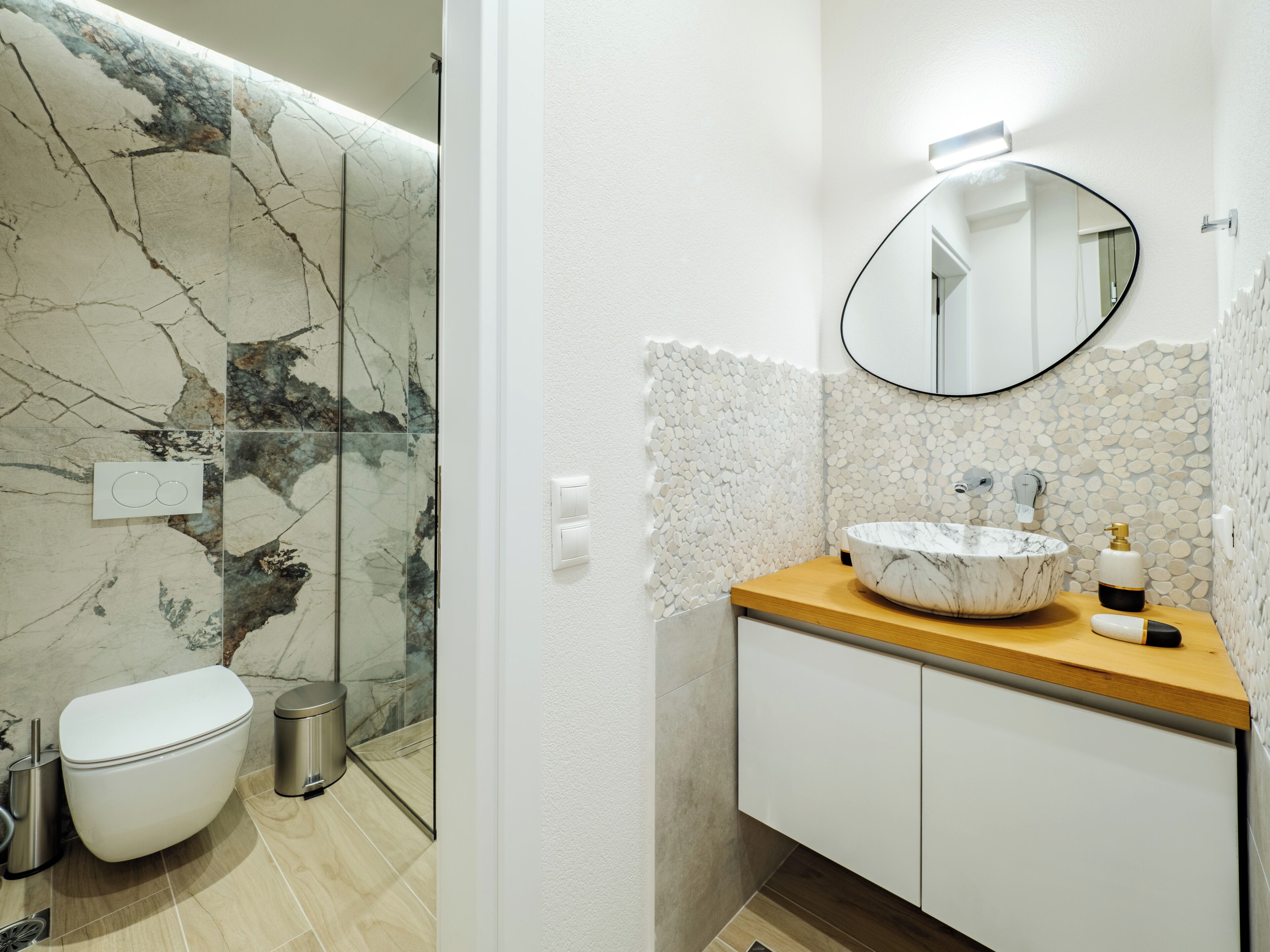 A bathroom with a marble wall, a round mirror, a wooden vanity, and a toilet.