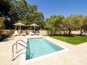 Ein Swimmingpool mit Liegestühlen und Sonnenschirmen, umgeben von einer Steinmauer und Grünpflanzen.