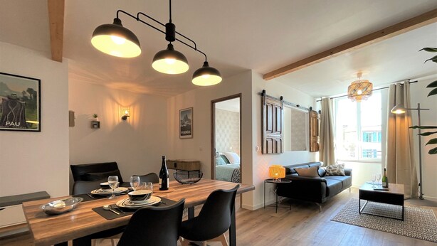 A modern living room with a dining area, featuring a wooden dining table, black chairs, and a sofa. There are pendant lights above the table and a floor lamp in the living room.