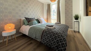 A modern bedroom with a geometric patterned wallpaper, a bed with a geometric patterned bedspread, a bedside table with a lamp, and a window with curtains.