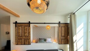 A modern living room with a black sofa, patterned cushions, a hanging woven light fixture, and a large mirror reflecting a bedroom with patterned wallpaper and a bed.