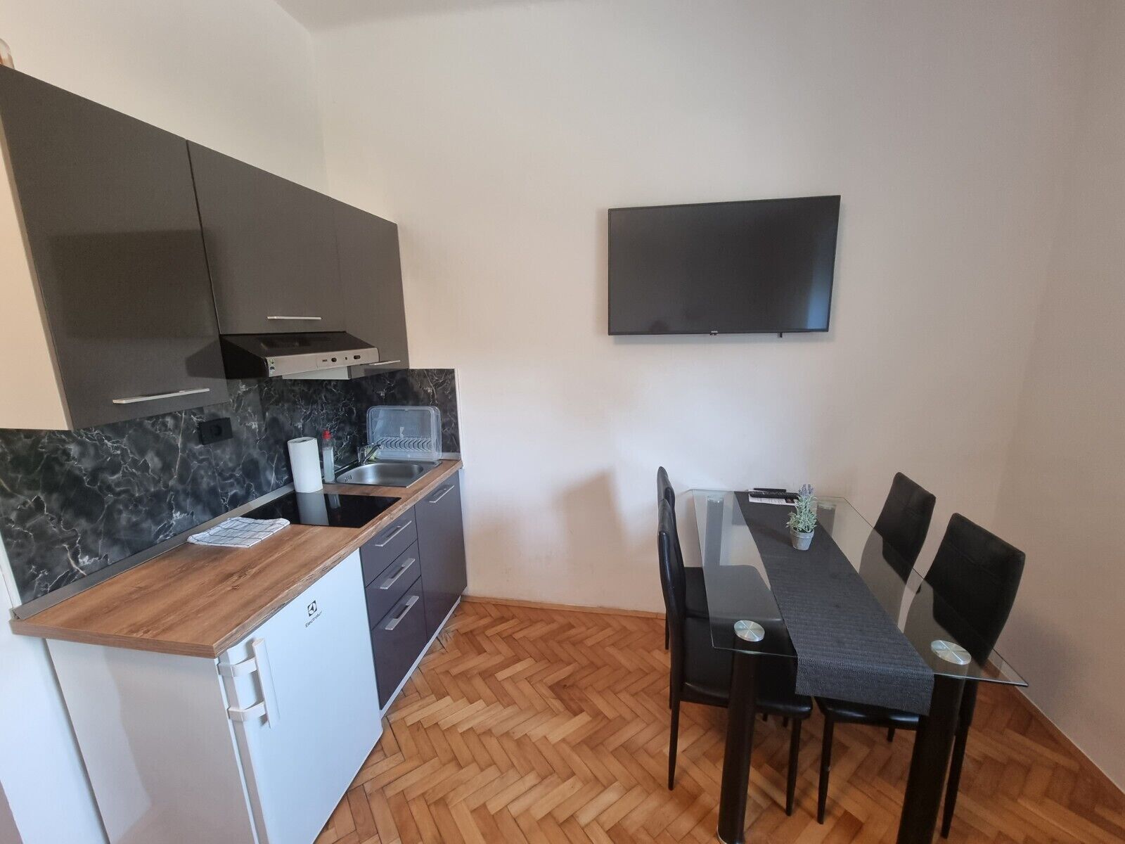 Una cocina compacta con horno integrado, microondas y lavamanos, una mesa de comedor con sillas y un televisor de pantalla plana montado en la pared.