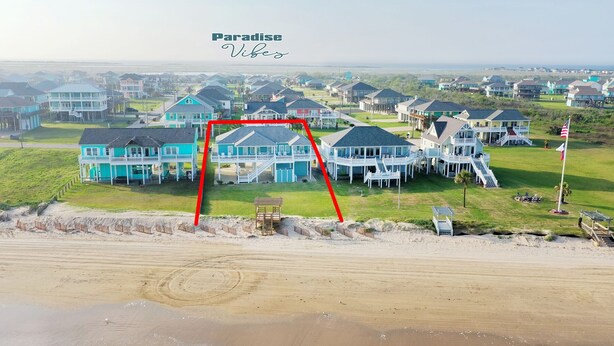 A coastal residential area with houses featuring blue exteriors and white balconies, a sandy beach in the foreground, and a clear view of the ocean in the background. - Paradise Vibes! 4-bedroom house in welcoming Bolivar Peninsula! (Bolivar Peninsula)