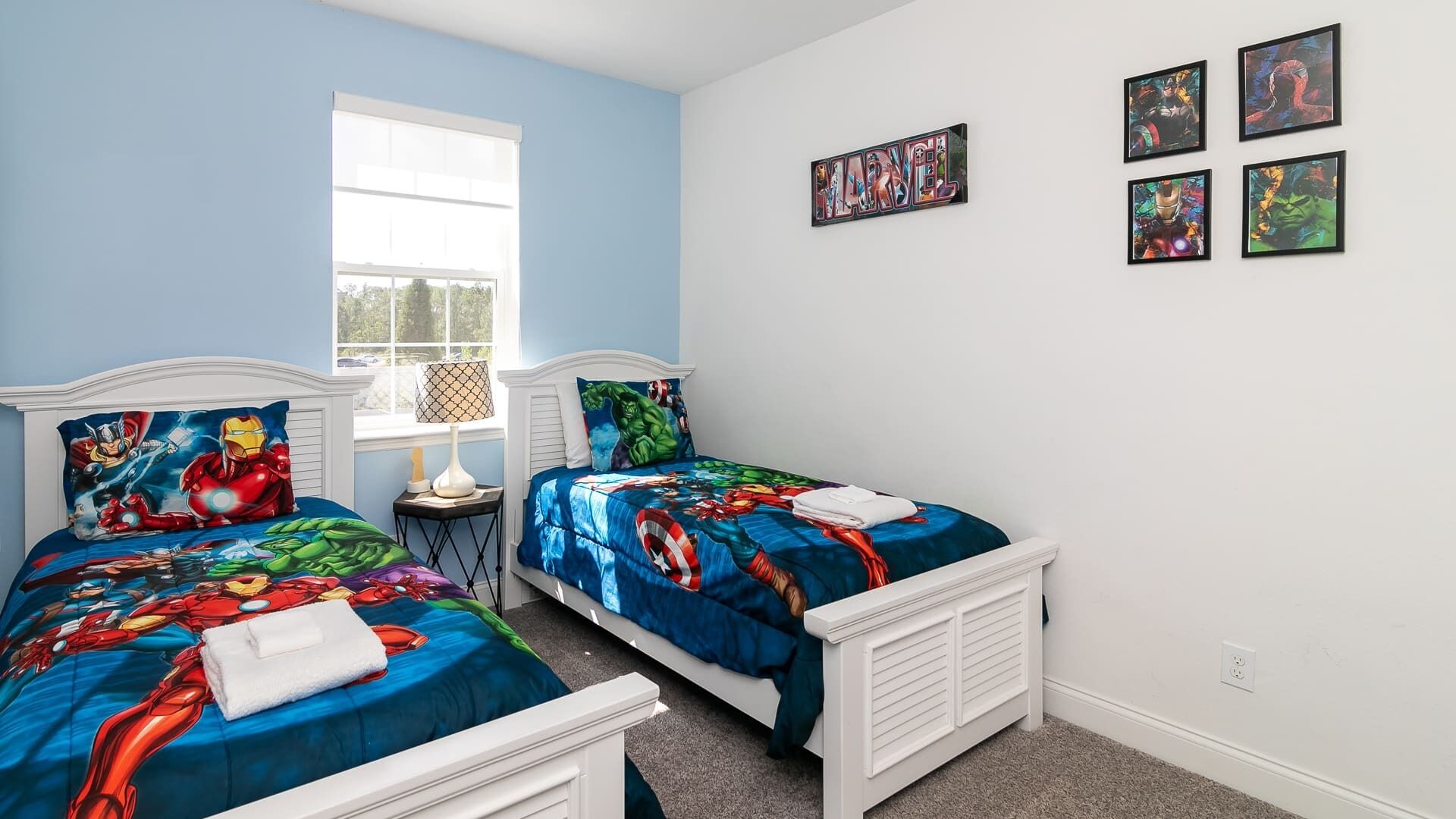 A bedroom with two beds, each with superhero-themed bedding, a window, and wall art.