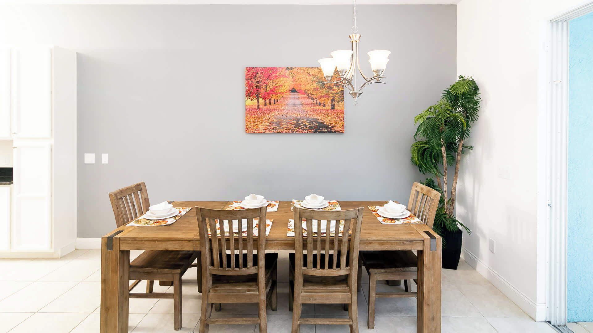 Un coin repas avec une table en bois dressée pour quatre personnes, un lustre et un tableau représentant un chemin bordé d’arbres.