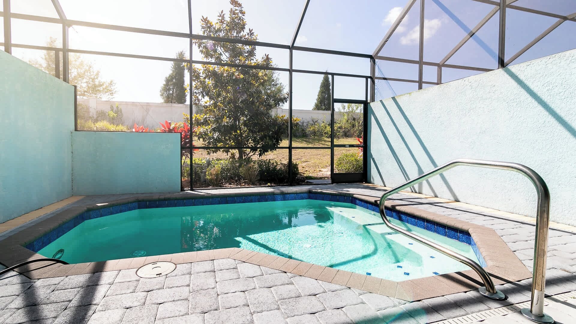 A swimming pool with a glass enclosure, a metal ladder, and a view of a garden and trees.
