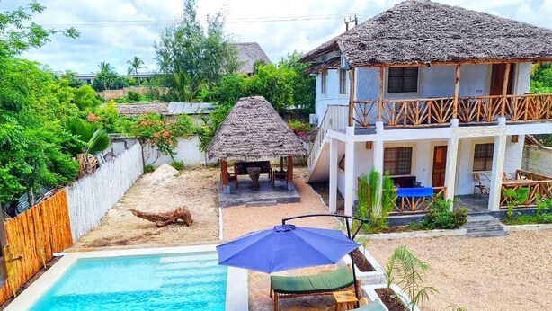 Une piscine entourée de chaises longues et d’un parasol, le tout délimité par une clôture en bois, avec une maison à toit de chaume à proximité.