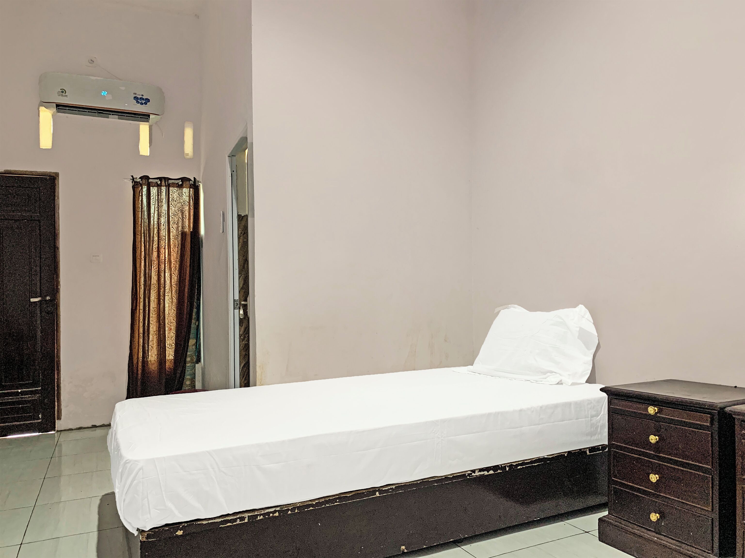 Un letto singolo con materasso e cuscino bianchi, una cassettiera in legno scuro e una finestra coperta da tende a motivi.