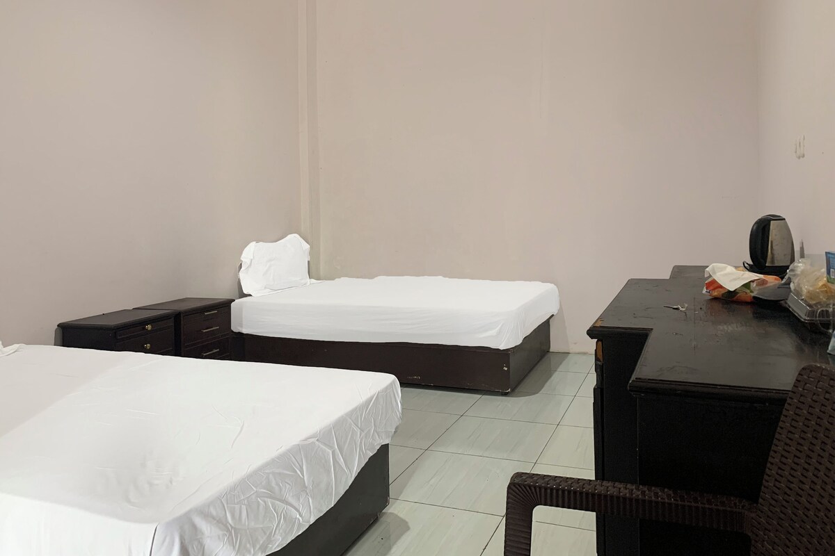 A hotel room with two beds, a desk with a chair, and a small table with a kettle and food items.