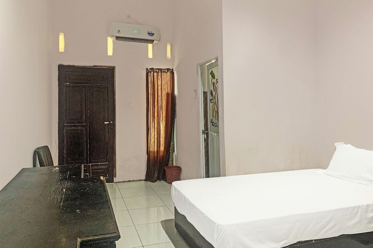 A hotel room with a bed, a desk, an air conditioner, a door, and a curtain.