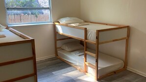 A bunk bed with a wooden frame and white bedding, a small desk with a chair, and a window with a view of greenery and a fence. - Eco-Shared Hype  (Miami)
