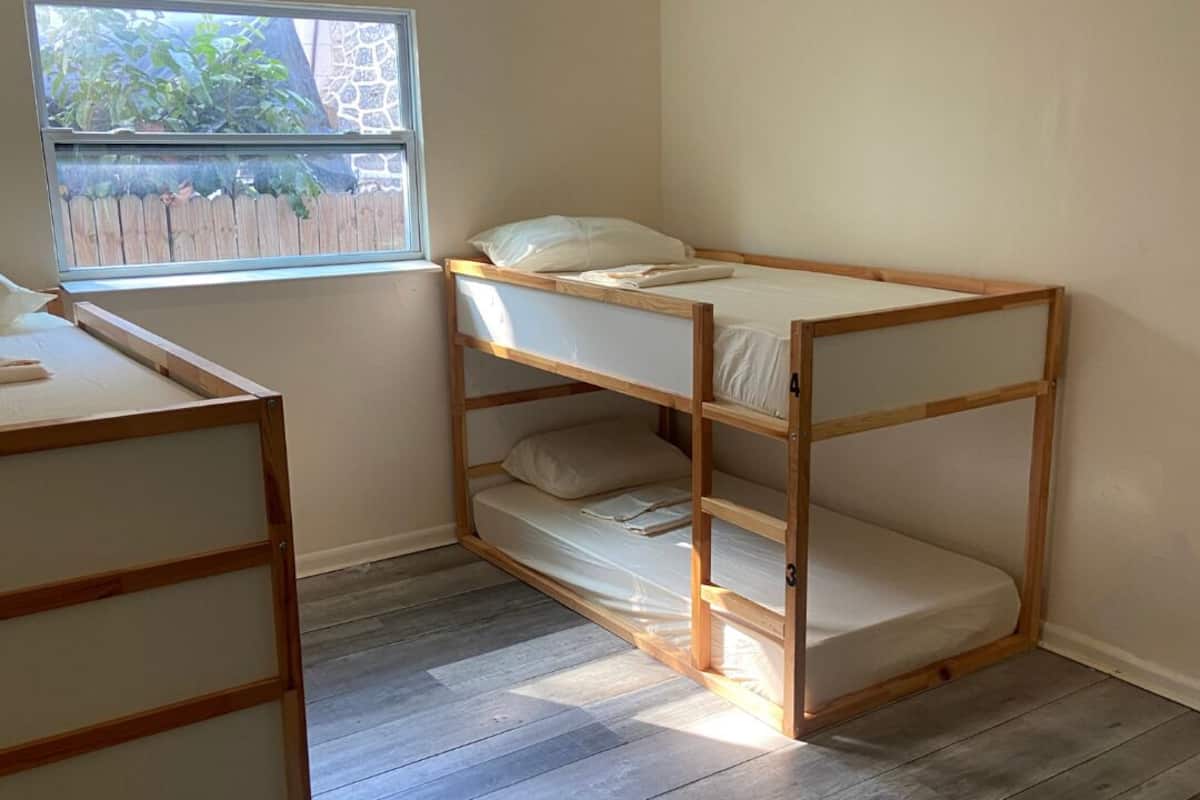 A bunk bed with a wooden frame and white bedding, a small desk with a chair, and a window with a view of greenery and a fence.