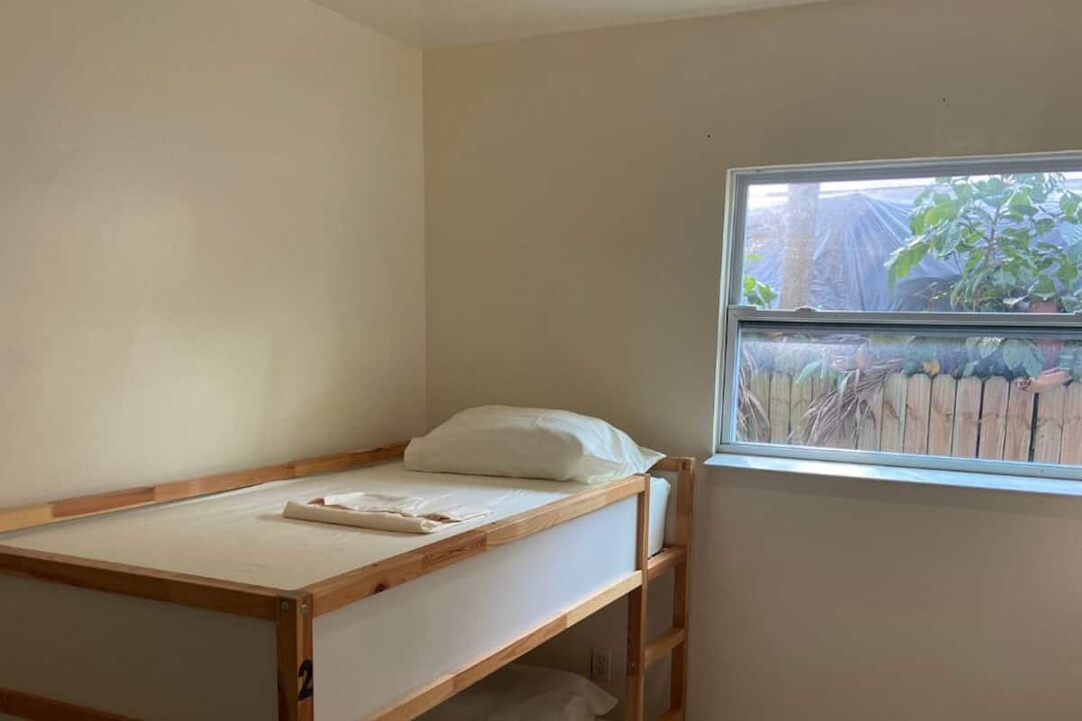 A bunk bed with a wooden frame and white bedding, a window with a view of a wooden fence and greenery, and a light-colored wall.