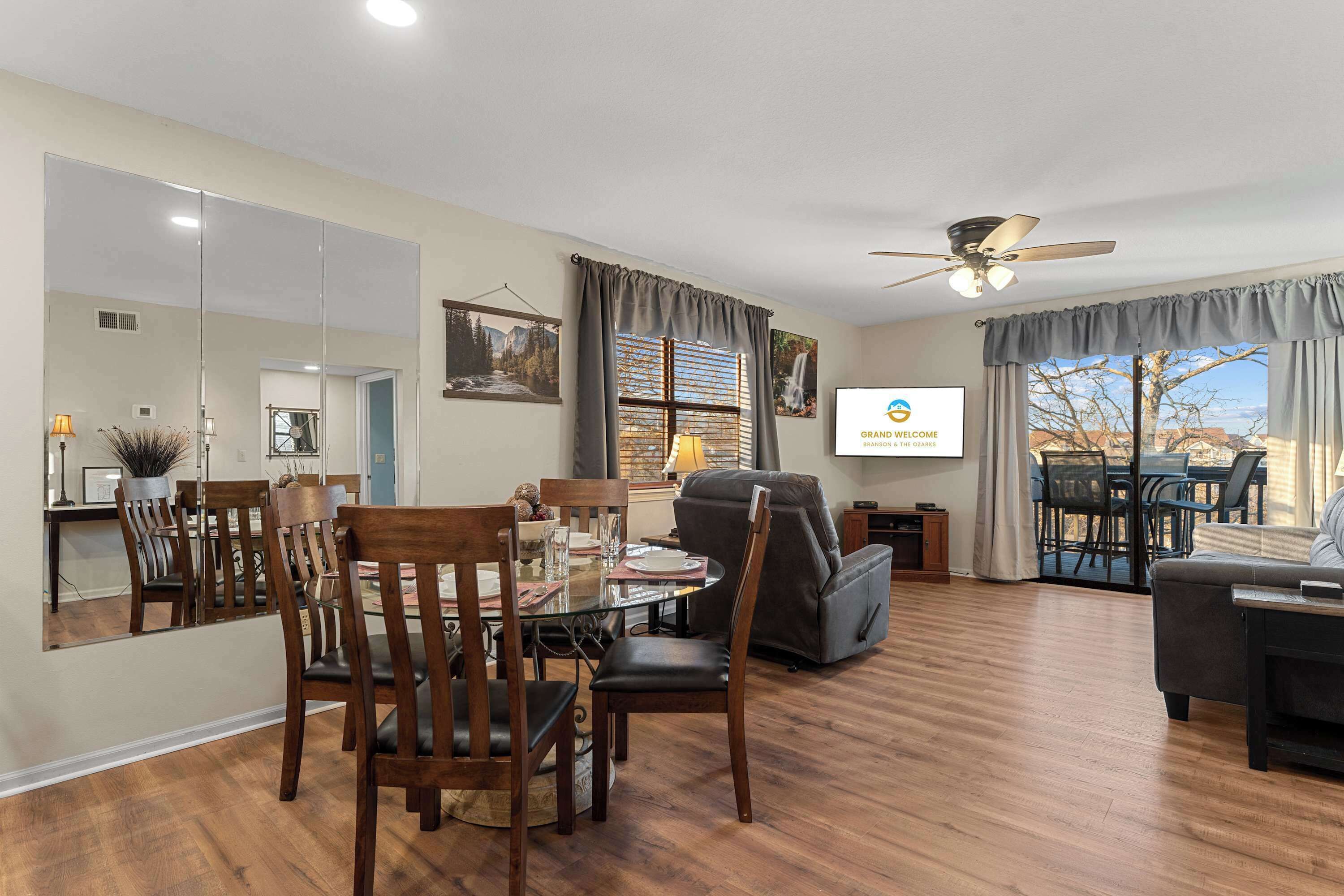 A spacious living room with a dining area, wooden flooring, a ceiling fan, and a large window with curtains.
