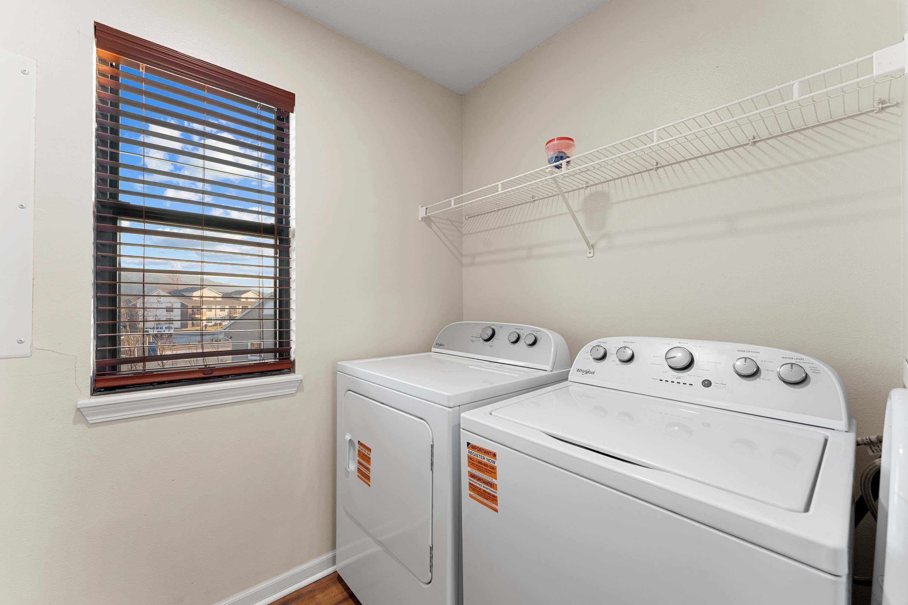 A laundry room with two white washers and dryers, a window with blinds, and a metal drying rack on the wall.