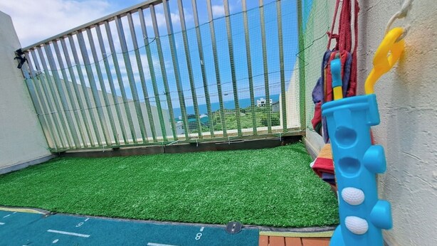 A children's play area with artificial grass, a play mat with numbers, a blue toy train, and a fence with a view of the sea. - Stayfarmsvill (Jeju City)
