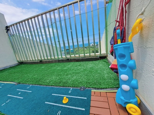 A children's play area with artificial grass, a play mat with numbers, a blue toy train, and a fence with a view of the sea.