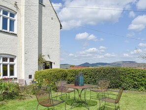A white building with a large window, a hedge, and a table with chairs in the foreground.