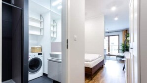 Un appartement moderne et compact comprenant un espace buanderie, une petite kitchenette et une chambre avec un lit et une table de chevet.