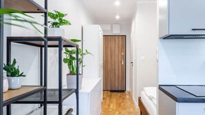 Une chambre d’hôtel compacte avec un plancher en bois, un lit, une kitchenette et une petite étagère ornée de plantes en pot.