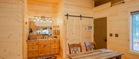 Interior de una cabaña de madera con comedor, cocina y un amplio ventanal.