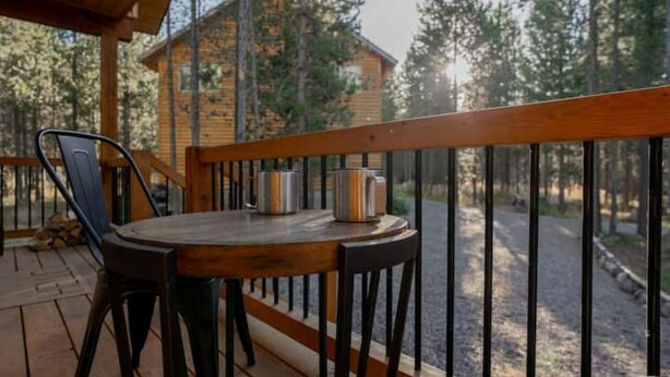 A wooden deck with a table and chairs, overlooking a forested area with a cabin in the background. - Tiny Timber-28 miles to Yellowstone (Island Park)