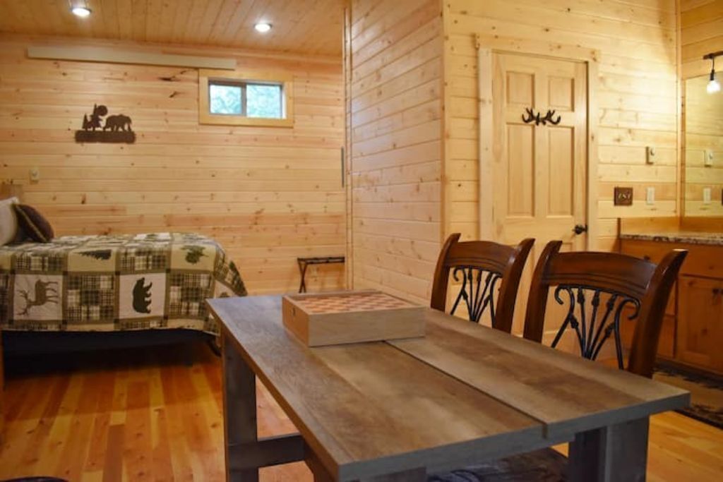 Interior acogedor de una cabaña con suelo de madera, una mesa de comedor con sillas, una cama con colcha, un ventanal pequeño y una puerta con adornos de asta de ciervo.