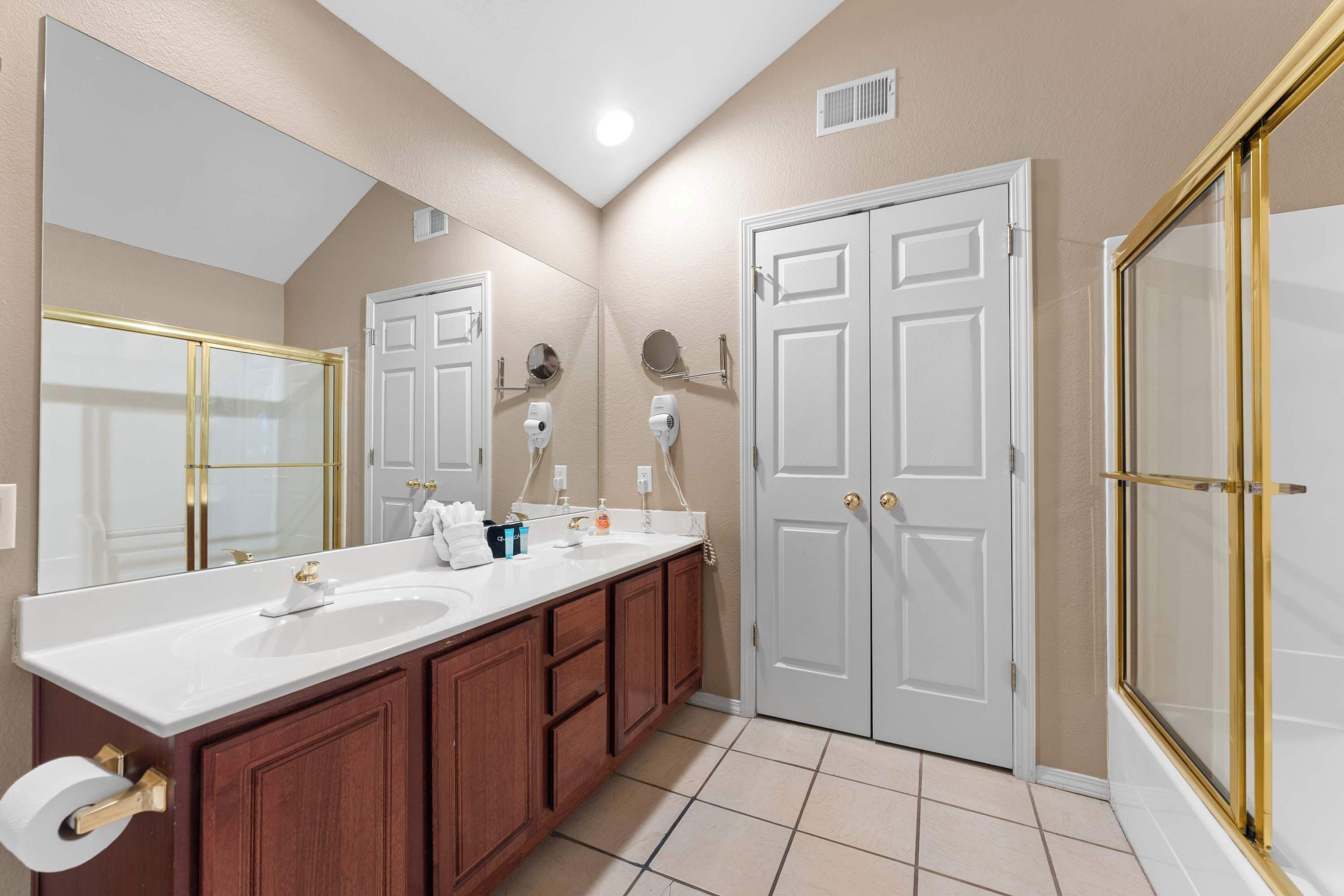 A bathroom with a large mirror, two sinks, wooden cabinets, and a shower area.