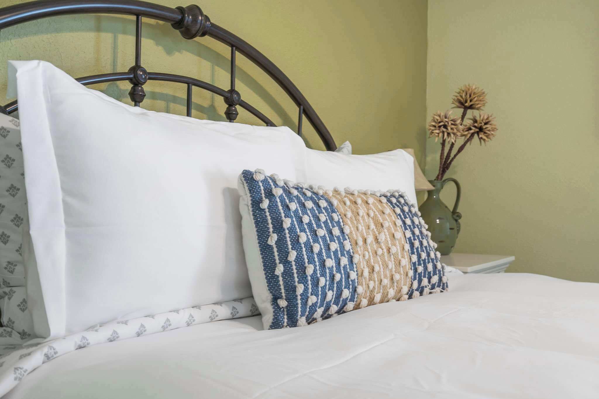 Una cama con ropa blanca y una almohada estampada, un cabecero de metal y un florero con flores secas sobre una mesita de noche.