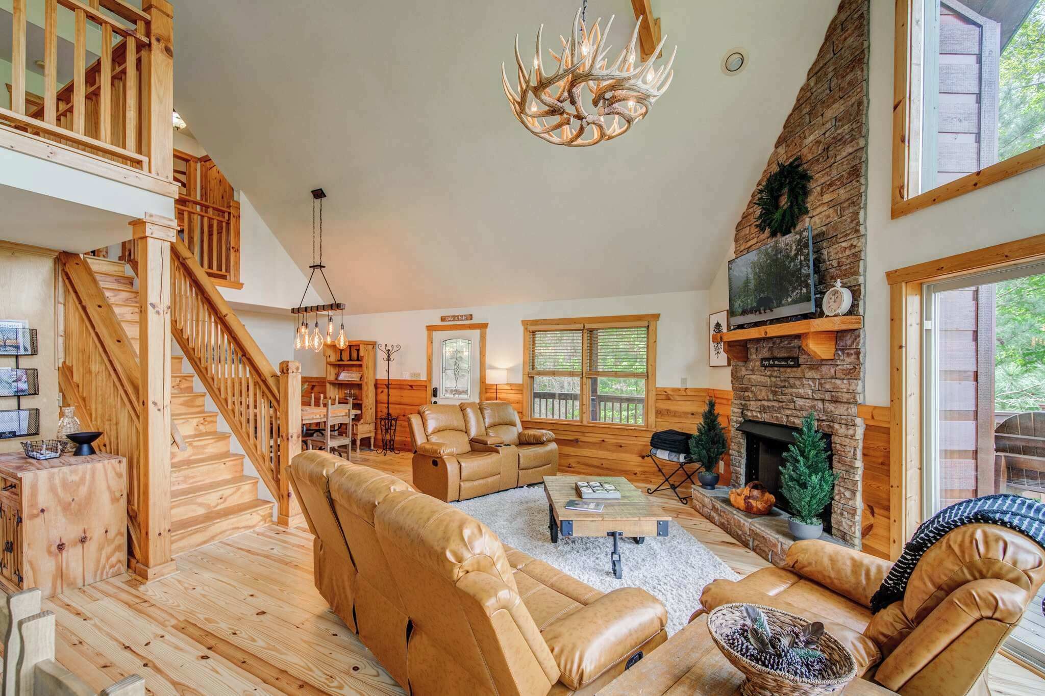 A spacious living room with a fireplace, leather sofas, a staircase, and a large chandelier.