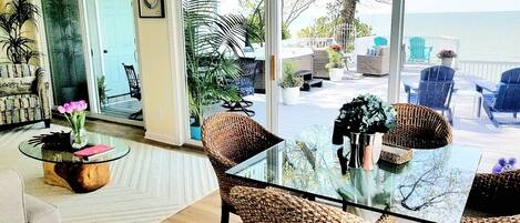 Un salon avec une table en verre, des fauteuils en osier, un canapé et une vue sur l’océan.