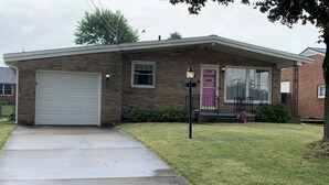 A single-story brick house with a white garage door, a small front porch with a pink door, and a well-maintained lawn. - Family Home, Blocks from Pro Football Hall of Fame, Fenced Yard, Pet Friendly (Canton)