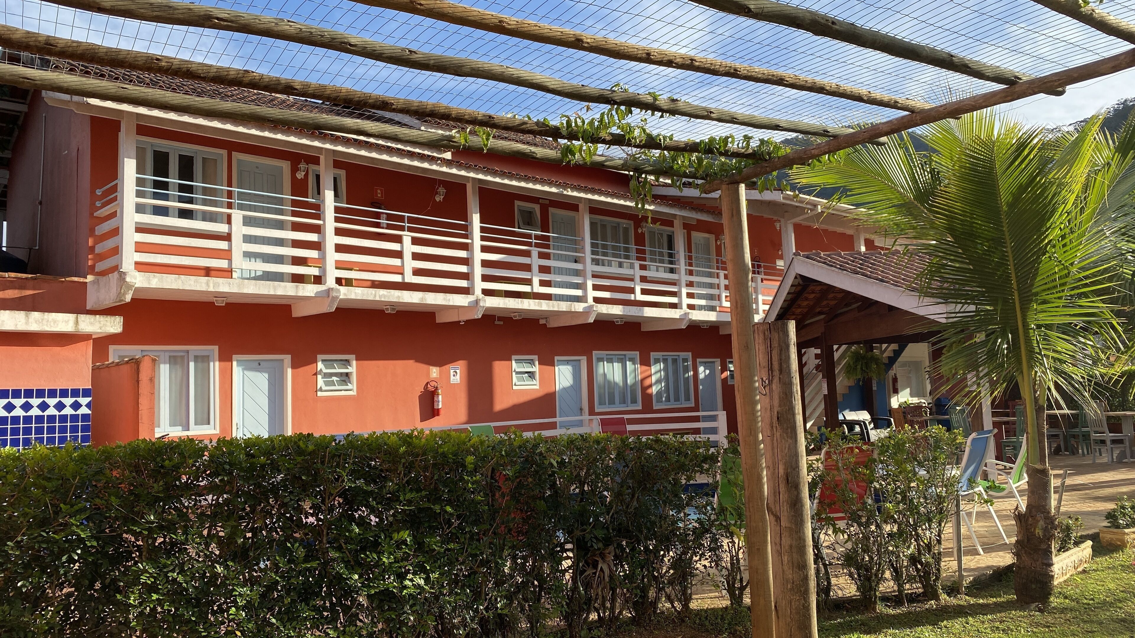 A red building with white balconies, a covered outdoor area, and a palm tree.