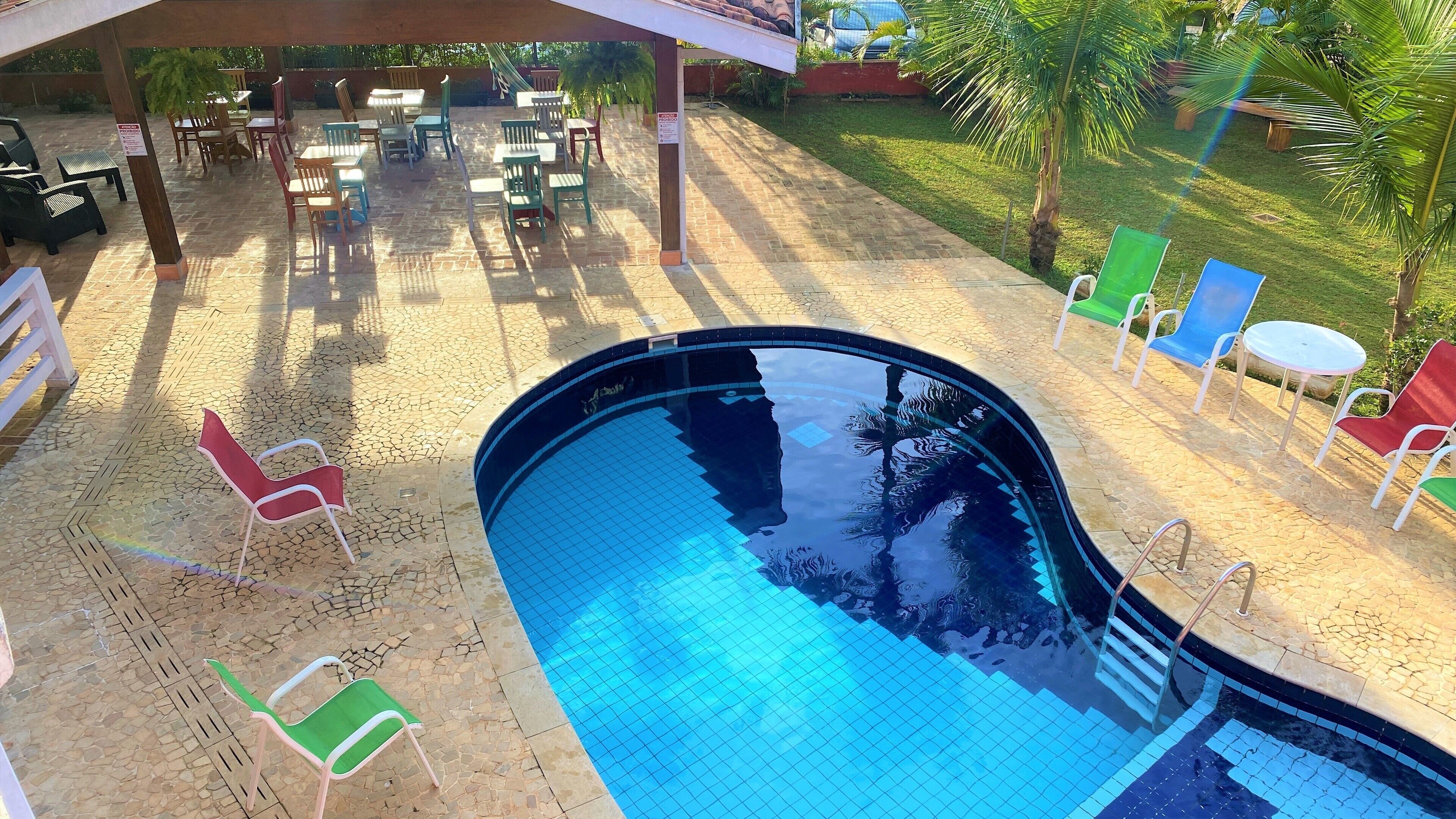 Una piscina con bordes azulejados, rodeada de zonas de estar al aire libre con sillas y mesas, y una sección cubierta con sombrillas.