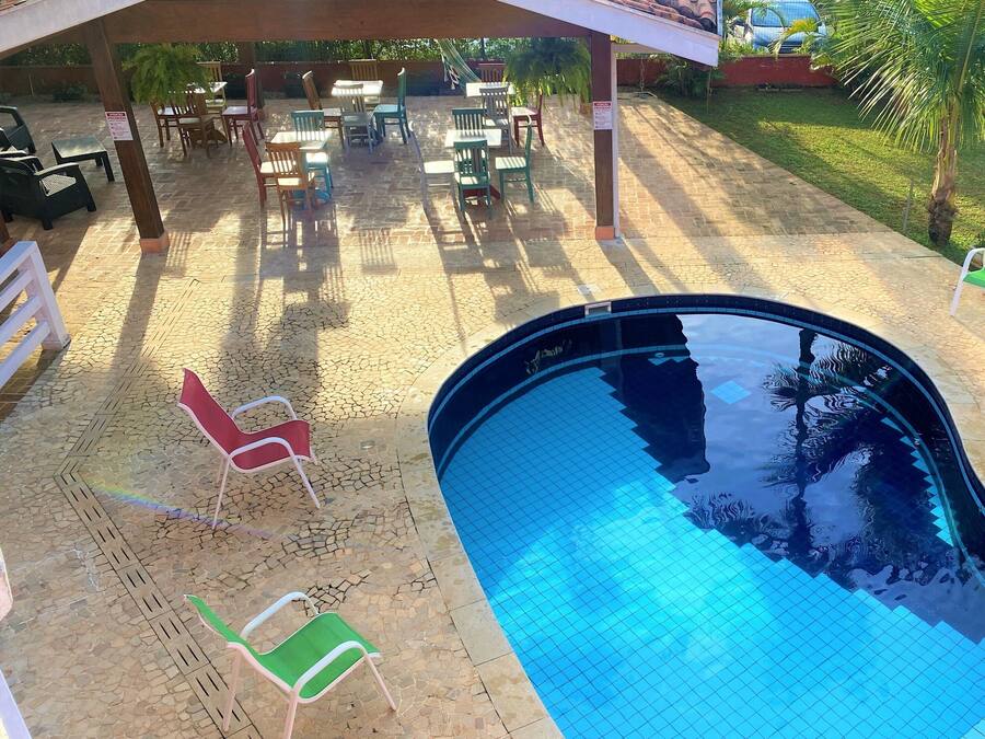 Una piscina con bordes azulejados, rodeada de zonas de estar al aire libre con sillas y mesas, y una sección cubierta con sombrillas.