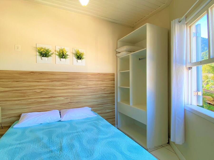 Quarto com cama, cabeceira de madeira, janela com vista e três plantas em vasos na parede.