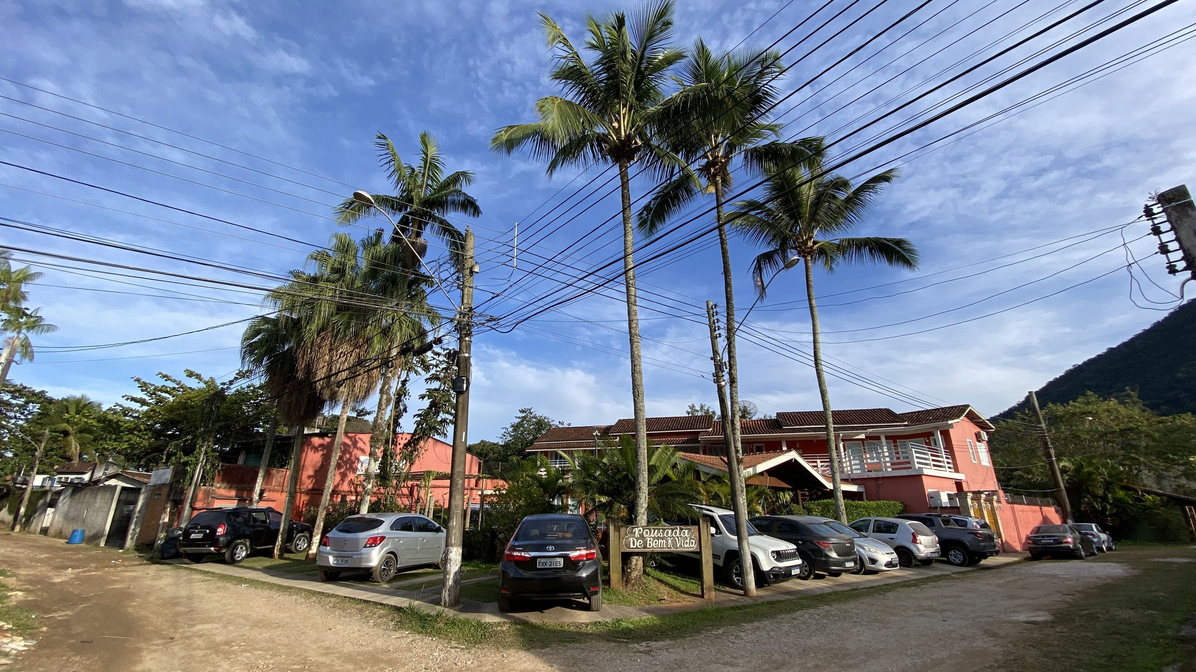 Una calle residencial con coches aparcados, palmeras y un edificio rojo con un cartel que dice 'Pousada De Bem K Vida'.