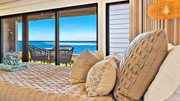 A bedroom with a large bed, multiple pillows, a view of the ocean, and a wicker lamp.