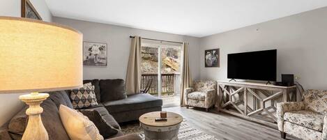 A living room with a sofa, armchairs, a coffee table, a TV, and a large window with curtains.