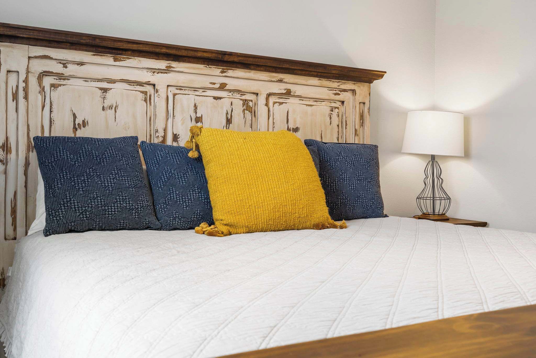 Een bed met een wit geborduurde sprei, een houten hoofdbord met verweerde verf en drie kussens in blauw en geel.