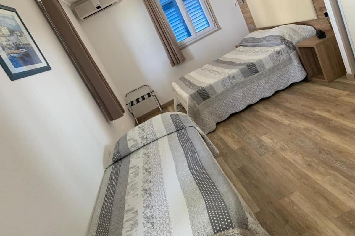 Ein Schlafzimmer mit zwei Einzelbetten, Holzboden, einem Fenster mit Jalousien, einer Wand-Klimaanlage und einem gerahmten Bild.