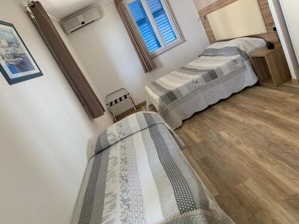 A bedroom with two single beds, a wooden floor, a window with blinds, a wall-mounted air conditioner, and a framed picture. - Hotel Le Bellevue (Saint-Florent)