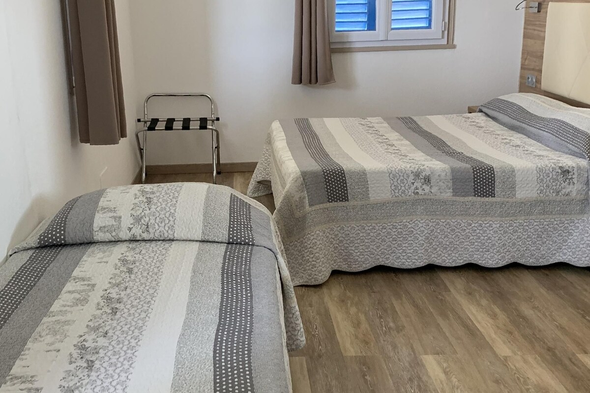 Una habitación pequeña y limpia con dos camas, una ventana con persianas, un perchero para toallas y piso de madera.