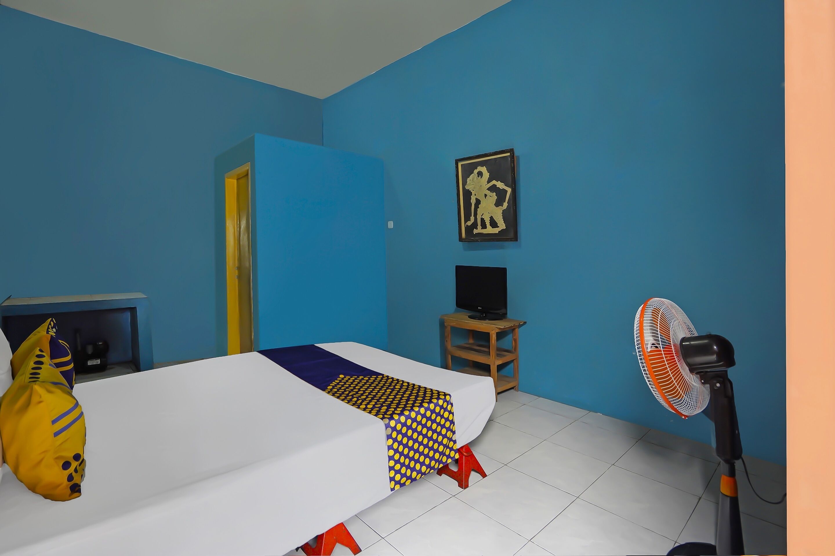 Une pièce avec un lit, un téléviseur posé sur un meuble, un ventilateur et un tableau accroché au mur.