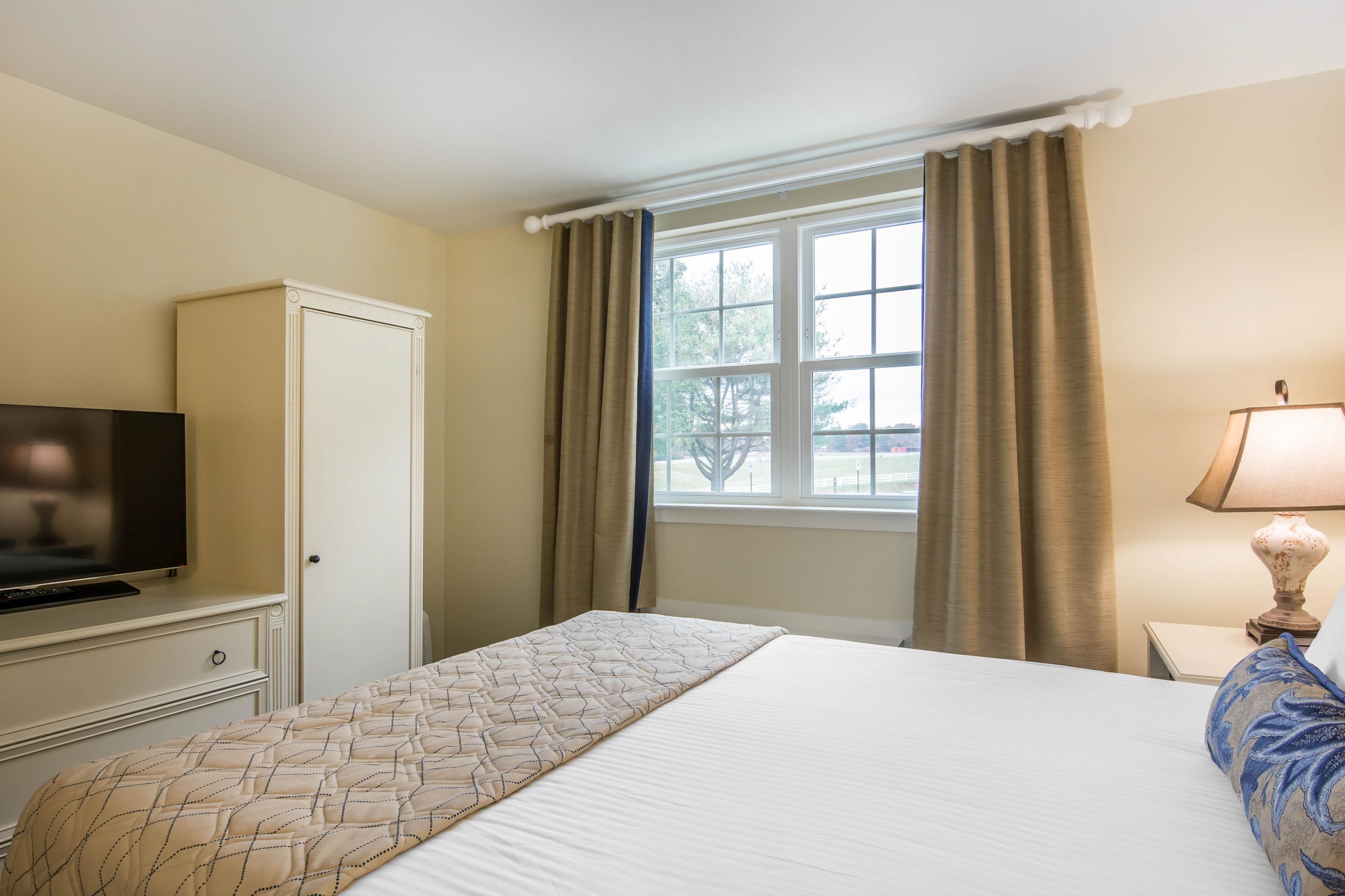 A hotel room with a bed, a television, a cabinet, a window with curtains, and a lamp.