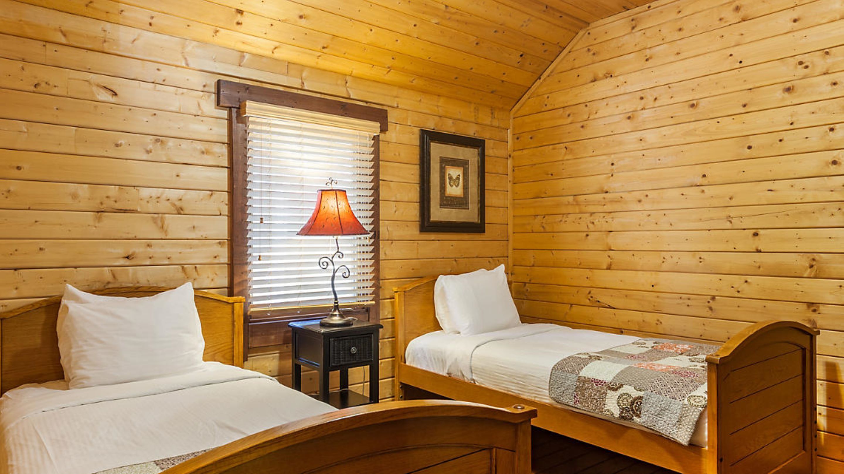 A wooden cabin room with two single beds, a nightstand with a lamp, a window with blinds, and a framed picture on the wall.