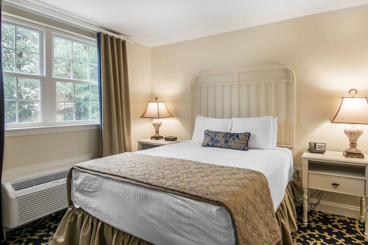 A hotel room with a large bed, two bedside tables with lamps, a window with curtains, and a radiator.