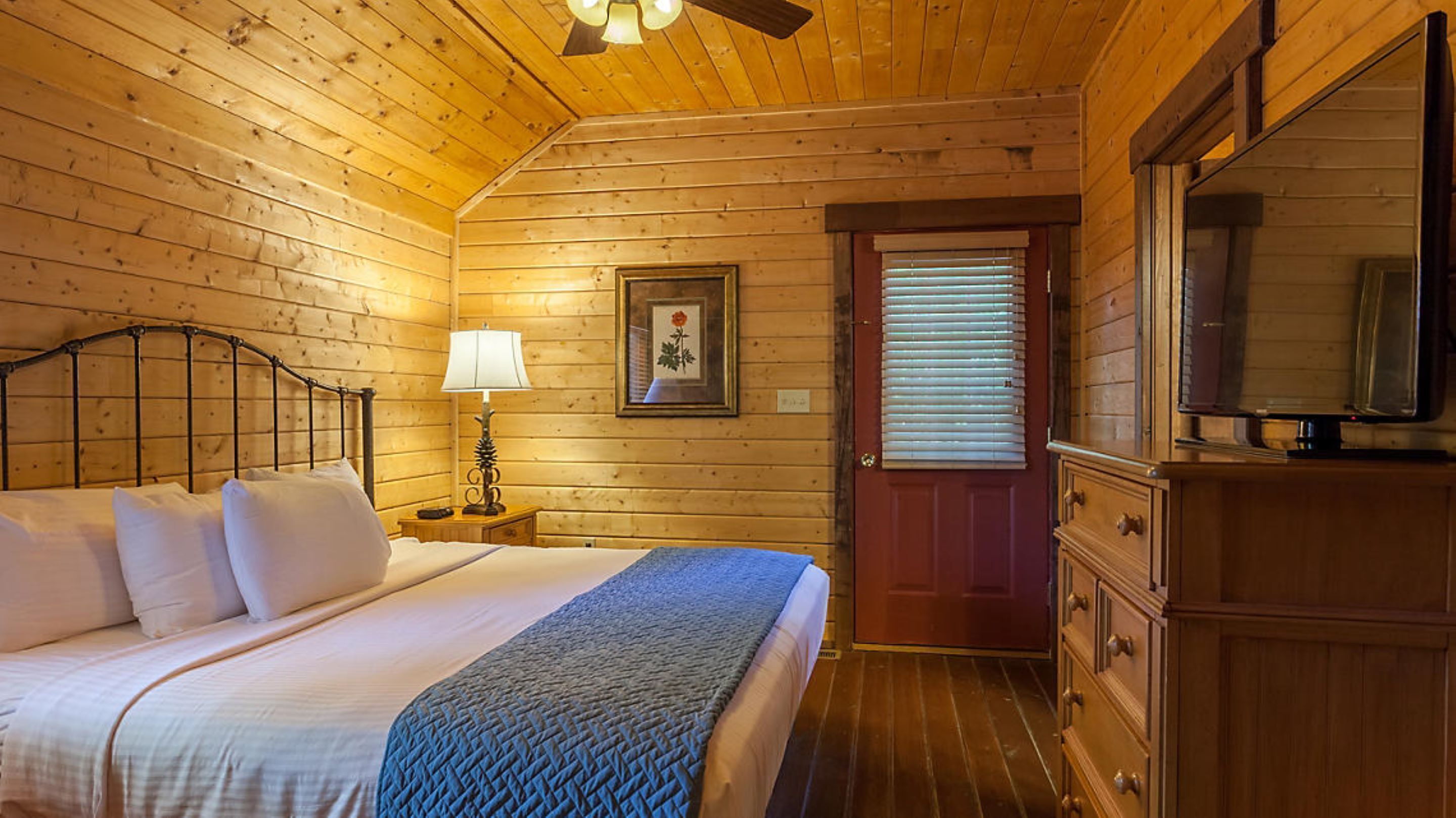 A wooden cabin bedroom with a bed, bedside lamp, dresser, and a television.
